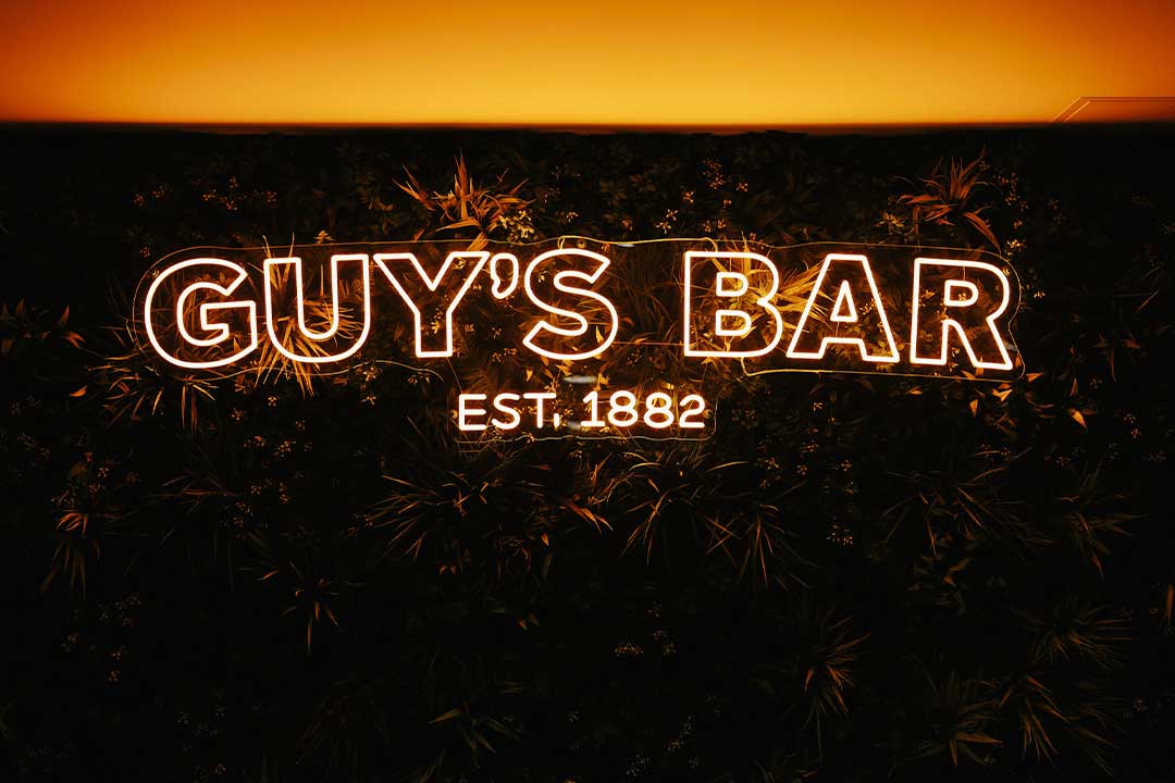 A wall with a forest design and an orange neon sign of the Guy's Bar logo mounted on it