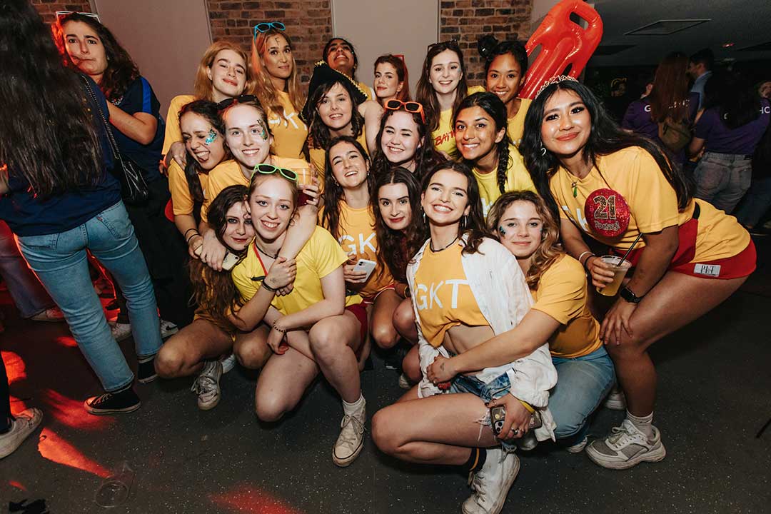 A group of students all crouched on the floor wearing glitter on their faces, a few of them have neon sunglasses on their head